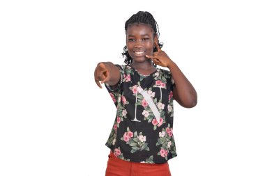 Çiçekli gecelikli genç bir kız beyaz arka planda dikiliyor ve parmağını göstererek telefon hareketinde bulunuyor..