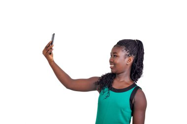 Örgülü genç bir kız beyaz arka planda durup gülerken kendini cep telefonuyla fotoğraflıyor..