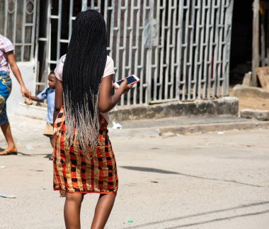 Abidjan, Fildişi Sahili - 14 Mart 2022: uzun saçlı, cep telefonunu elinde tutan ve yolda yürüyen genç bir kadının portresi.