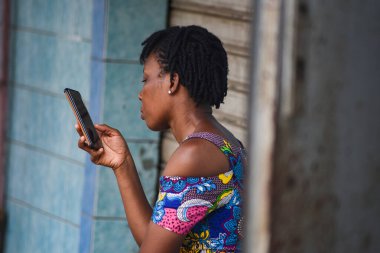 Abidjan, Fildişi Sahili - 19 Mart 2022: örgülü saçlı genç kadın duvarın yanında oturmuş cep telefonuna bakıyor..