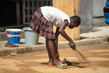 Abidjan, Ivory Coast - March 30, 2022: Young downcast girl sweeps her shop front with a traditional broom.