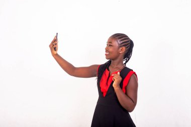 Kırmızı ve siyah elbiseli güzel Afrikalı bir kız beyaz arka planda durup cep telefonuyla gülümserken kendini fotoğraflıyor..
