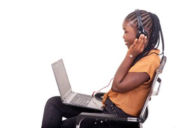 Pantolon giymiş genç bir bayan öğrenci evde sandalyede oturuyor ve kulaklıklarıyla bilgisayardan gülümsüyor..