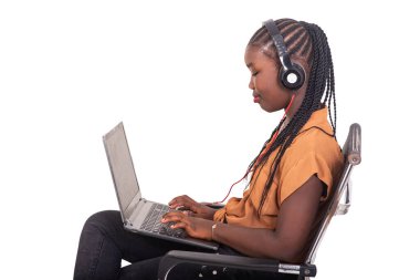 Pantolon giymiş genç bir bayan öğrenci evde sandalyede oturuyor ve kulaklıklarıyla bilgisayardan gülümsüyor..