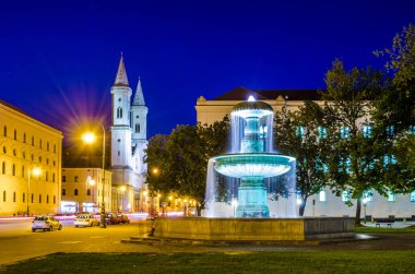 Münih 'teki üniversitenin önündeki fıskiyenin gece görüşü ve arka planda Aziz Ludwig Kilisesi.