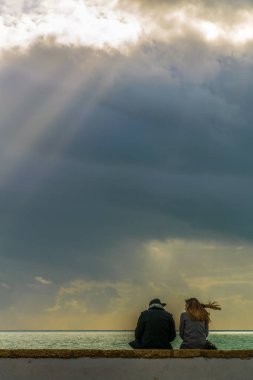 Genç bir çift deniz kenarındaki bir duvarda oturuyor ve yoğun bir bulutun içinden geçen güneş ışınlarını seyrediyor.