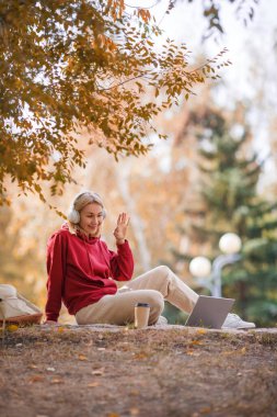 Görüntülü arama. Kulaklıklı gülümseyen kadın elinde kahveyle dizüstü bilgisayarda oturuyor ve sonbaharda halka açık parkta birini selamlıyor, dikey fotoğraf, seçilmiş odak..