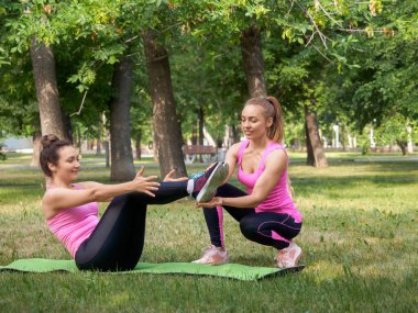 Açık hava takibi. Çekici beyaz kadın yaz mevsiminde kamu parkında arkadaşının yardımıyla tekne dengesi talimi yapıyor.