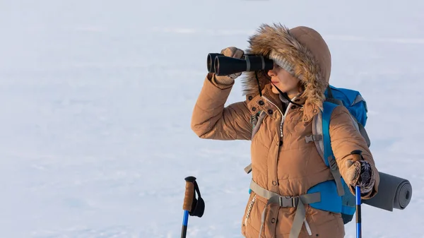 バックパックとスキーポール付きの黄色のフードダウンジャケットの女性は冬に雪の平原に立って双眼鏡を通して見て、腰を上げ、選択された焦点. — ストック写真
