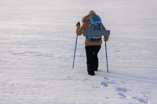 青いバックパックとスキーポールが雪に覆われた平野の葉の痕跡、バックビュー、選択された焦点を歩くとベージュダウンジャケットの人間. — ストック写真