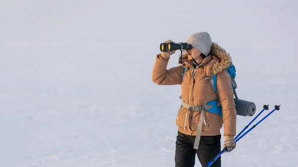 バックパックとスキーポールを手にダウンジャケットの女性は冬に雪の平原に立って双眼鏡を通して見て、選択された焦点. — ストック写真