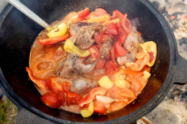 Shurpa in a cauldron at the stake. Tomato, red pepper and lamb soup cooked on hot coals in nature. Hot beef hunting goulash or bograch soup with paprika, small, vegetables and spices.