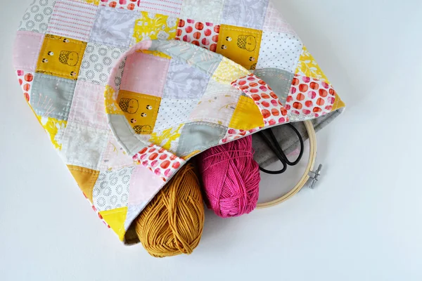 Patchwork tote bag with embroidery hoop, black scissors, pink and mustard yarn on white background