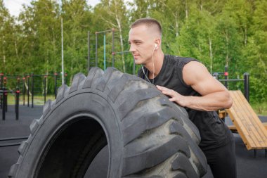 Muscular bearded tattooed fitness man moving large tire in street gym. strength exercise for lifting a tire by a young athlete