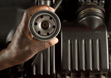South African or American hand holding oil filter with modern car engine background clipart