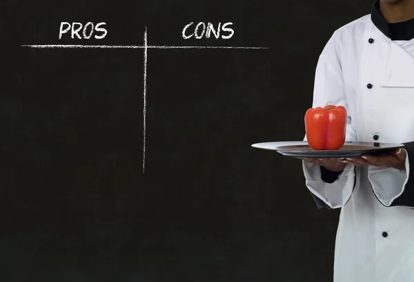 African american chef holding healthy food with chalk pros and cons on blackboard background — Stock Photo, Image