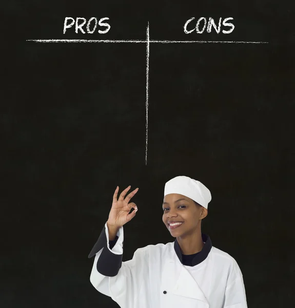 African american woman chef with chalk pros and cons on blackboard background — Stock Photo, Image