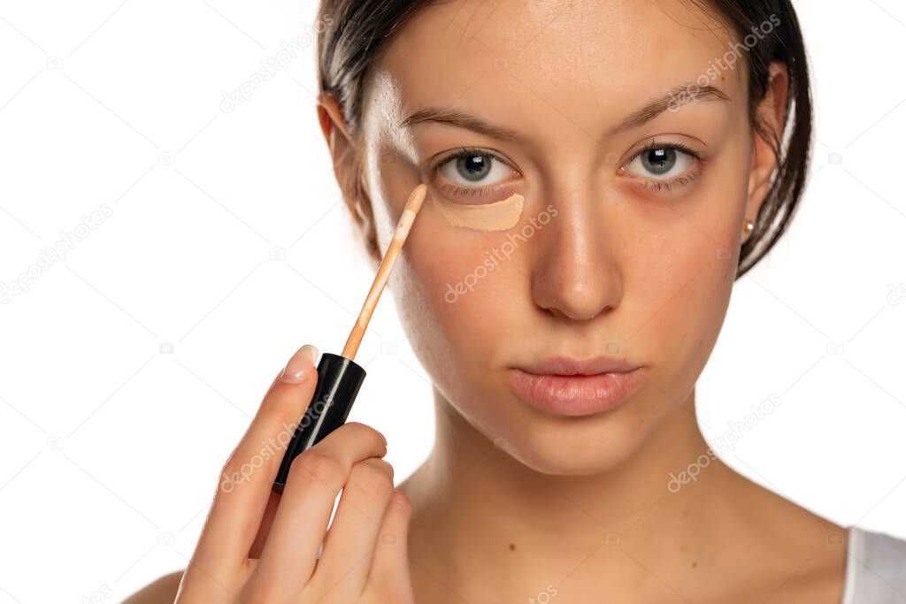Portrait of young woman with blue eyes applying concealer under her eye on a white background