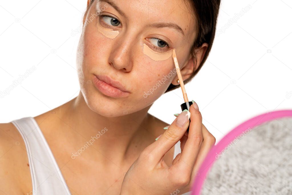 Portrait of young woman with blue eyes applying concealer under her eyes on a white background