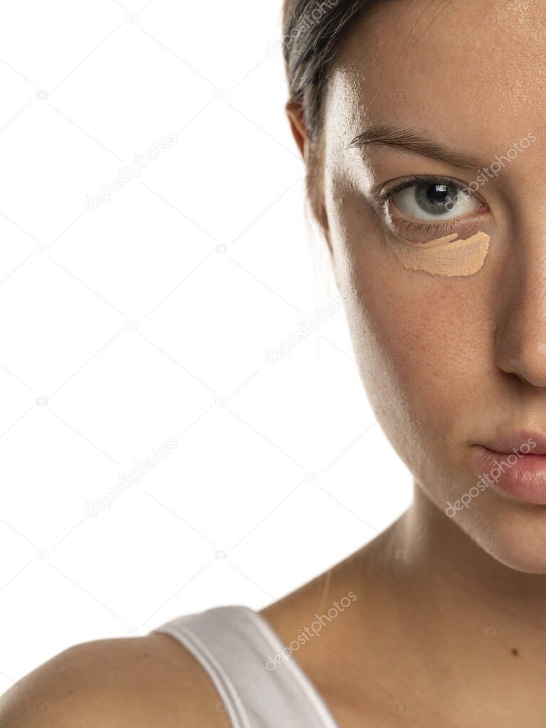 Closeup of young woman with blue eyes with concealer under her eye on a white background