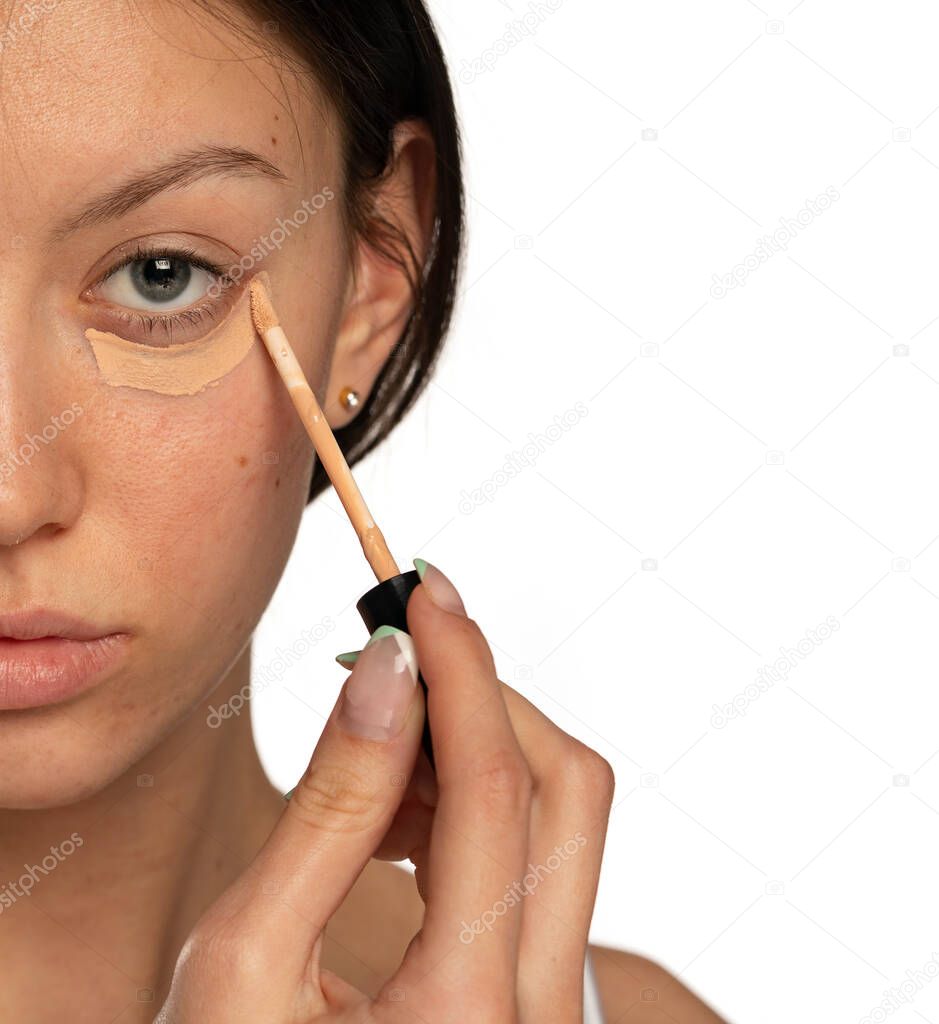 Closeup of young woman with blue eyes applying concealer under her eye on a white background