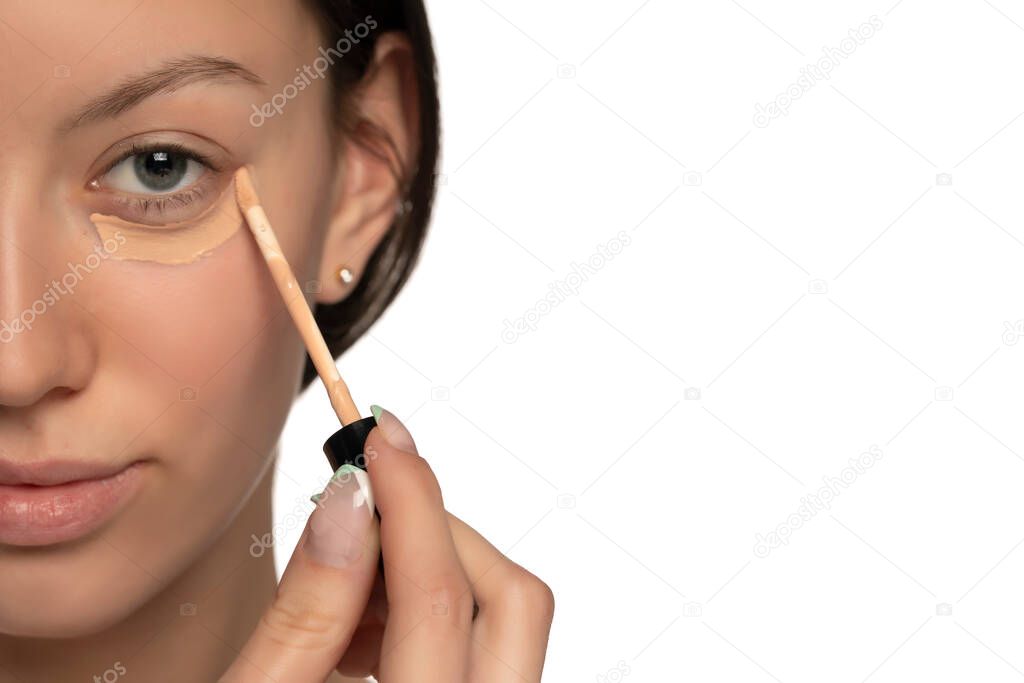 Closeup of young woman with blue eyes applying concealer under her eye on a white background