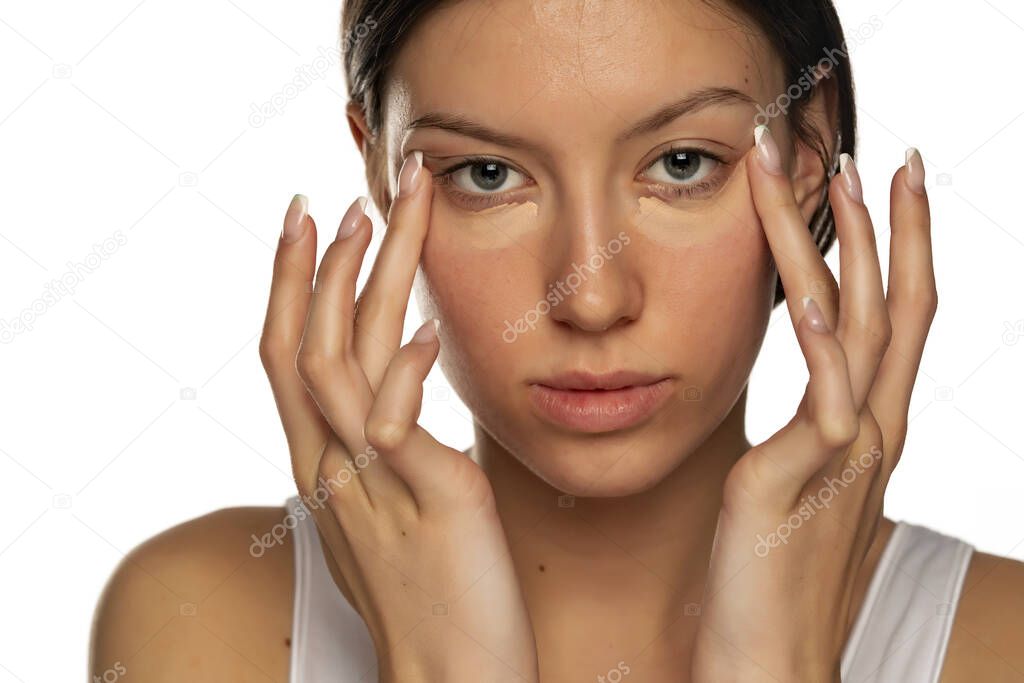 Portraitf young woman with blue eyes applying concealer under her eyes on a white background