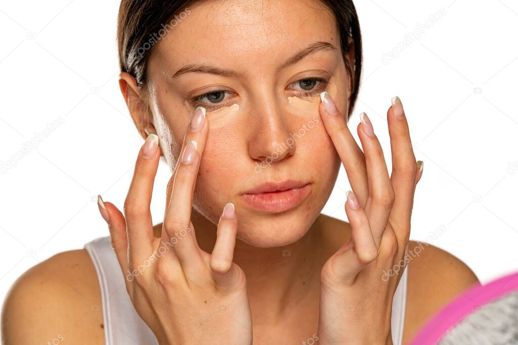 Portrait of young woman applying concealer under her eyes on a white background