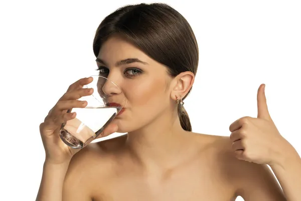 若い美しい女性がガラスから水を飲み白い背景に親指を立てて — ストック写真