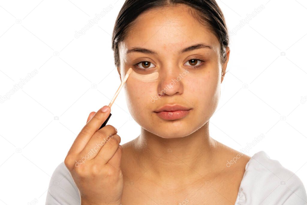 closeup of a young beautiful woman applies concealer under her eye on a white background