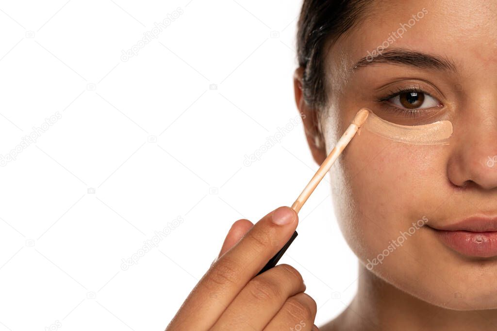 closeup of a young beautiful woman applies concealer under her eye on a white background