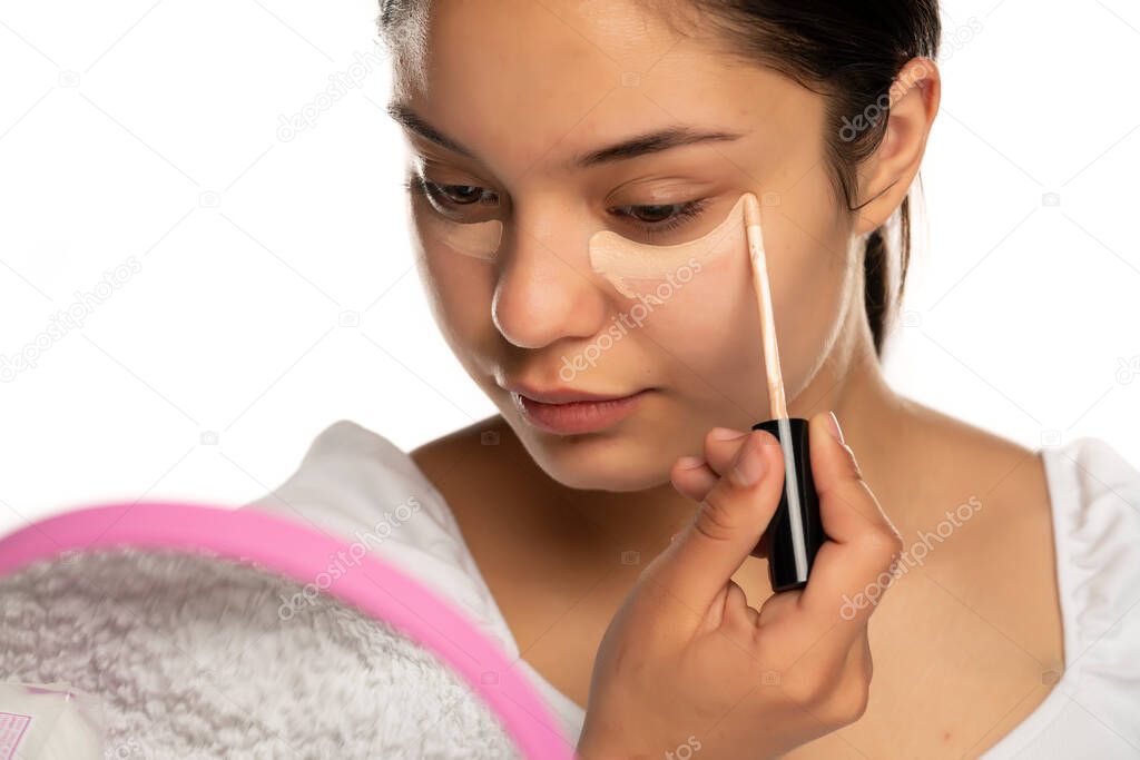 a young woman applies concealer under her eyes on a white background