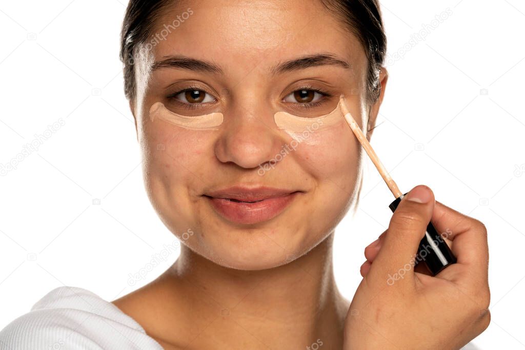 closeup of a young beautifu smilingl woman applies concealer under her eye on a white background