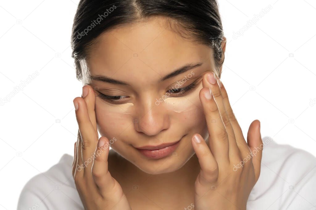 Head shot of a young beautiful woman applies concealer under her eyes on a white background