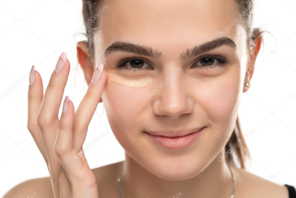 smiling teen girl applyes concealer under her eyes on white background