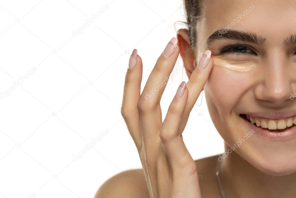 happy teen girl applyes concealer under her eyes on white background