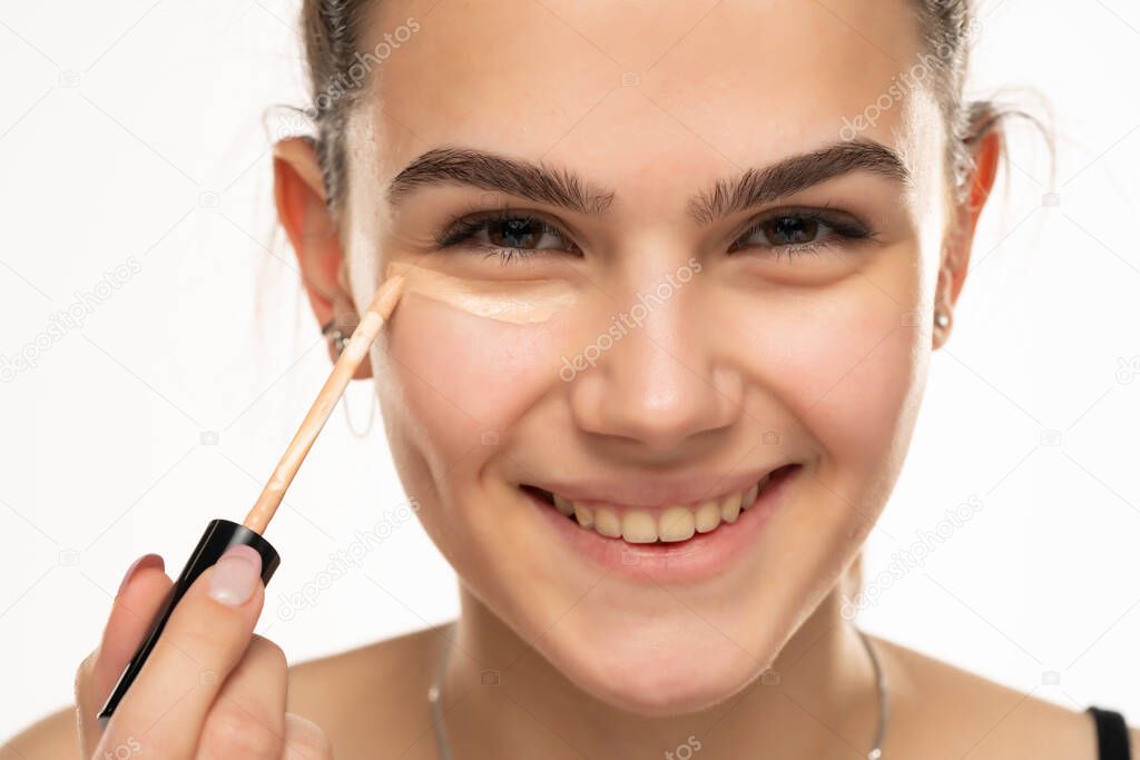 smiling teen girl applyes concealer under her eyes on white background