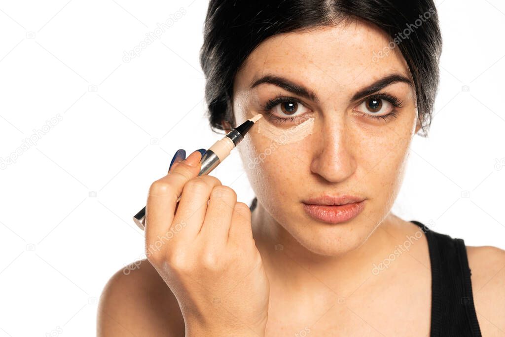 Portrait of young beautiful woman applying concealer with applicator on white background
