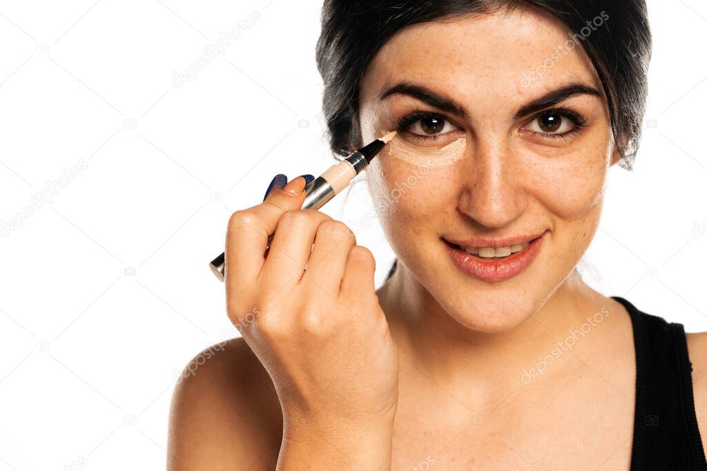 Portrait of young beautiful woman applying concealer with applicator on white background
