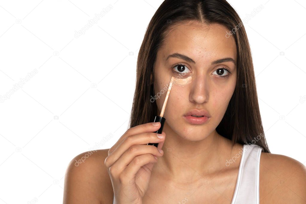 a young woman applies concealer under her eyes on a white background