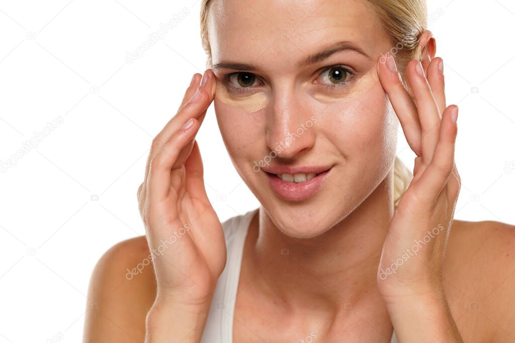 Young blond smiling woman applying concealer under her eyes on a white background