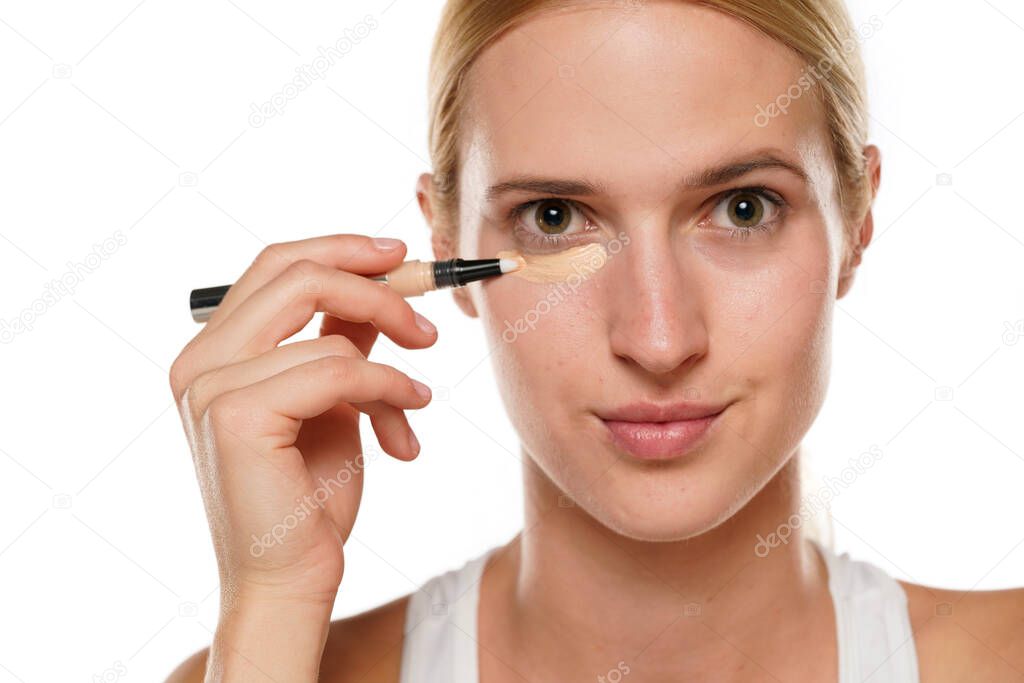 Young blond woman applying concealer under her eyes on a white background