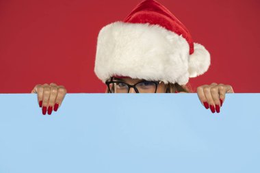 Noel, Noel, Noel Baba, insanlar, reklam, satış konsepti. Kırmızı arka planda boş mavi panonun arkasına bakan Noel Baba şapkalı mutlu kadın.