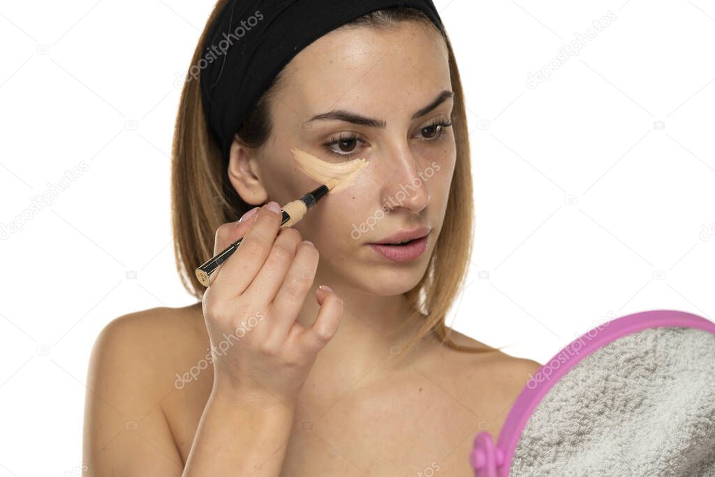 Young woman applyes concealer under her eyes on white background