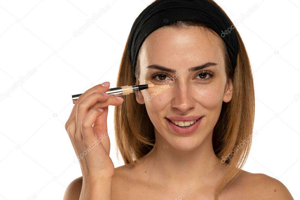 Young smiling woman applyes concealer under her eyes on white background