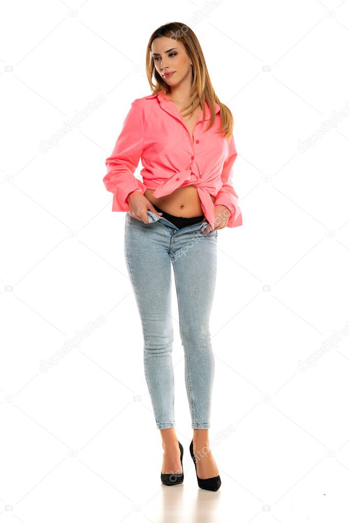 Charming young woman posing in studio in pink shirt and unbuttoned blue jeans on a white background
