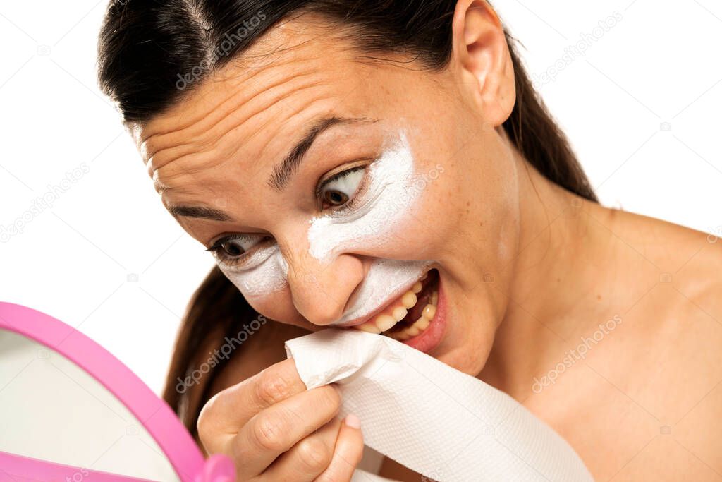 young woman with a cosmetic product under the eyes and nose cleaning her teeth with a wet wipe on a white background
