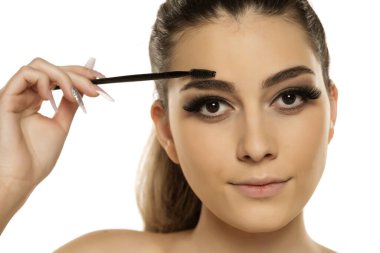 Genç bir kadın kaş şeklini fırçayla düzeltiyor, beyaz arkaplan