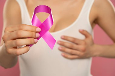 Kapalı tişörtlü genç kadın pembe arka planda kanser farkındalığı kurdelesi taşıyor.