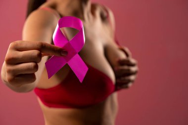 Kırmızı sutyenli genç bir kadın pembe arka planda kanser farkındalığı kurdelesi taşıyor.